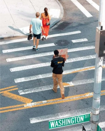 A delivery person carries stacked pizza boxes across a crosswalk in a city, while other pedestrians walk nearby on a sunny day.