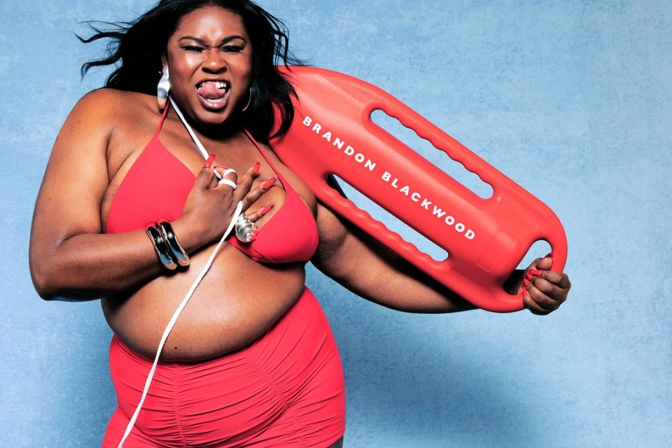 Adult female in red swimwear with a lifeguard rescue can with text 'Brandon Blackwood' against a light blue studio backdrop.