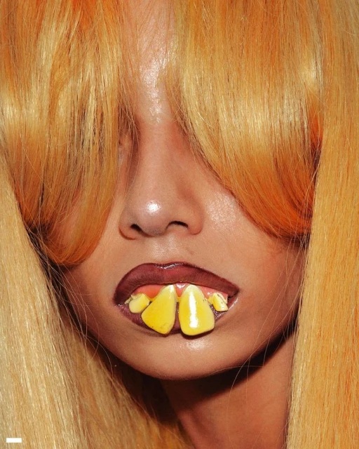 Close-up portrait of a person with blonde hair and yellow tooth coverings, in a studio lighting setting, focusing on face and mouth.