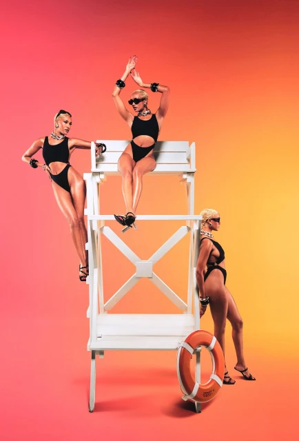 Three women in black swimwear pose with a lifeguard chair and ring buoy against a vibrant gradient background of red, orange, and yellow.