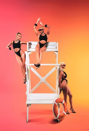 Three women in black swimwear pose with a lifeguard chair and ring buoy against a vibrant gradient background of red, orange, and yellow.