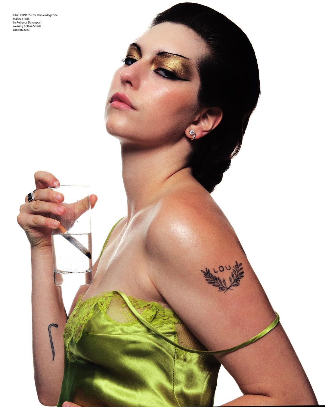 A woman in a green satin top holds a glass of water, featuring gold eyeshadow and a small shoulder tattoo, against a plain white background.