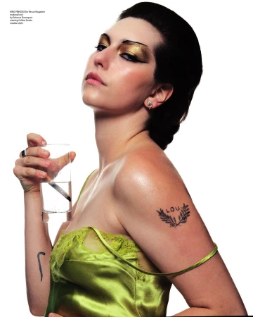 A woman in a green satin top holds a glass of water, featuring gold eyeshadow and a small shoulder tattoo, against a plain white background.