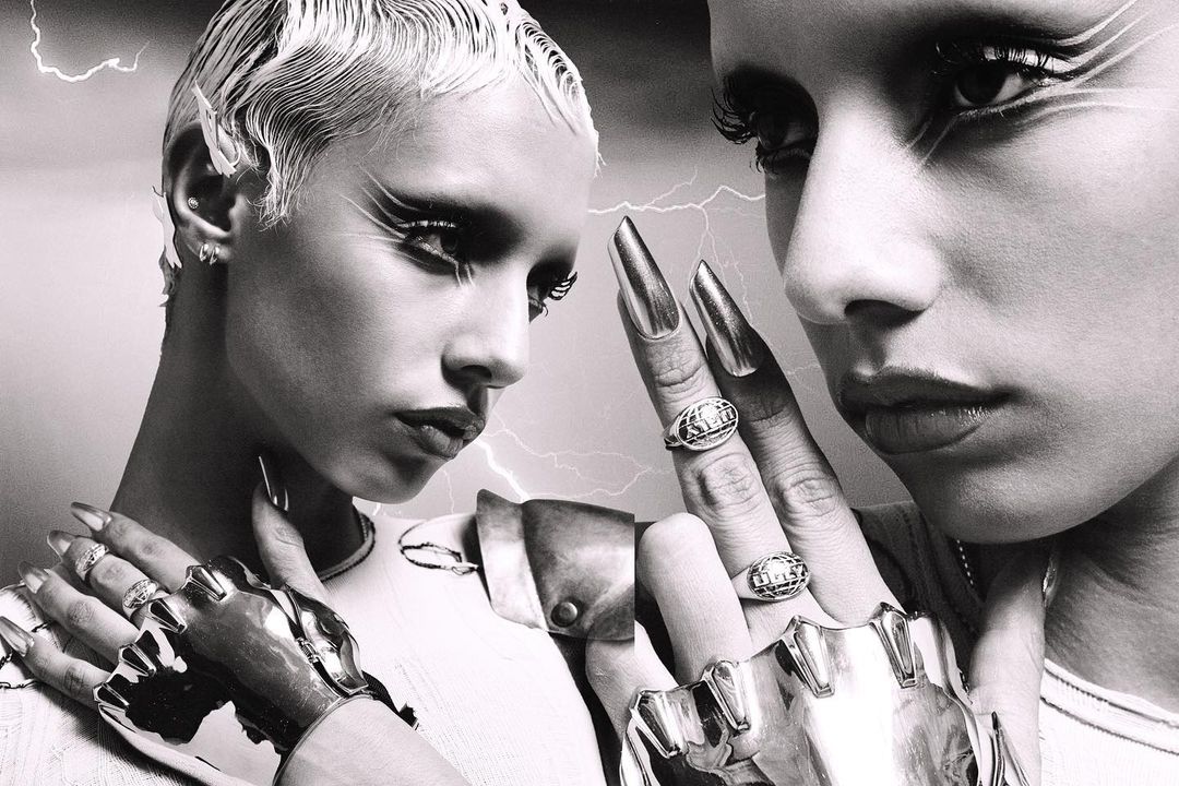 Monochrome portrait features two striking women with dramatic makeup, metallic nail extensions, and sculptural metal gauntlets on a lightning-streaked backdrop.