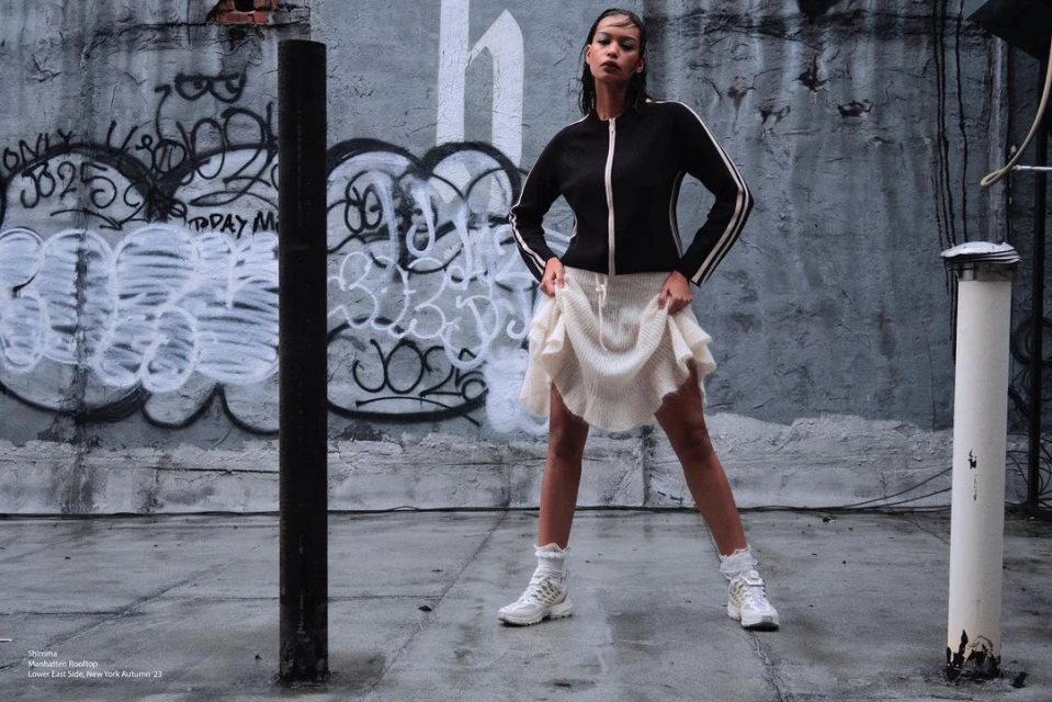 A woman poses confidently on a rooftop in a black jacket, white skirt, and white sneakers against a graffiti wall, bathed in soft light.