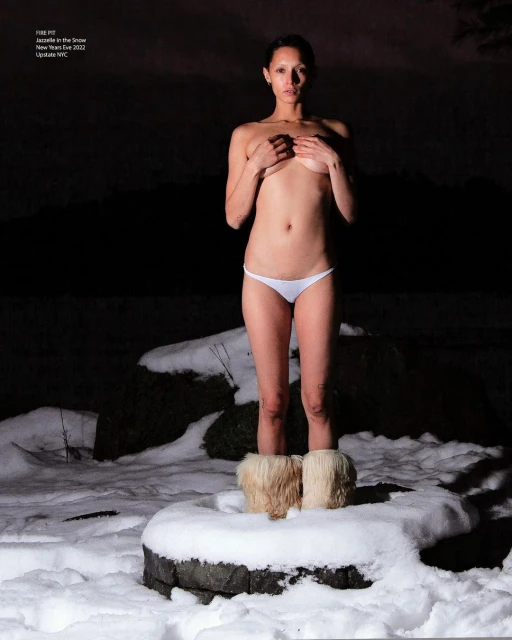Woman in underwear stands in snow, covering her breasts with hands, wearing furry boots, with a dark mountain background at night.