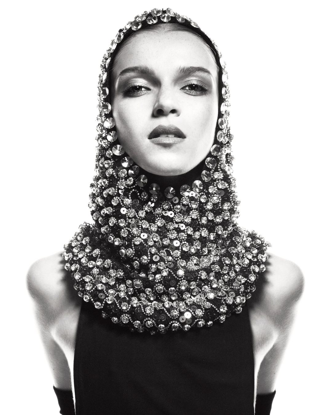 Monochrome fashion portrait of a woman wearing a beaded headpiece and sleeveless black top against a bright white backdrop in studio.