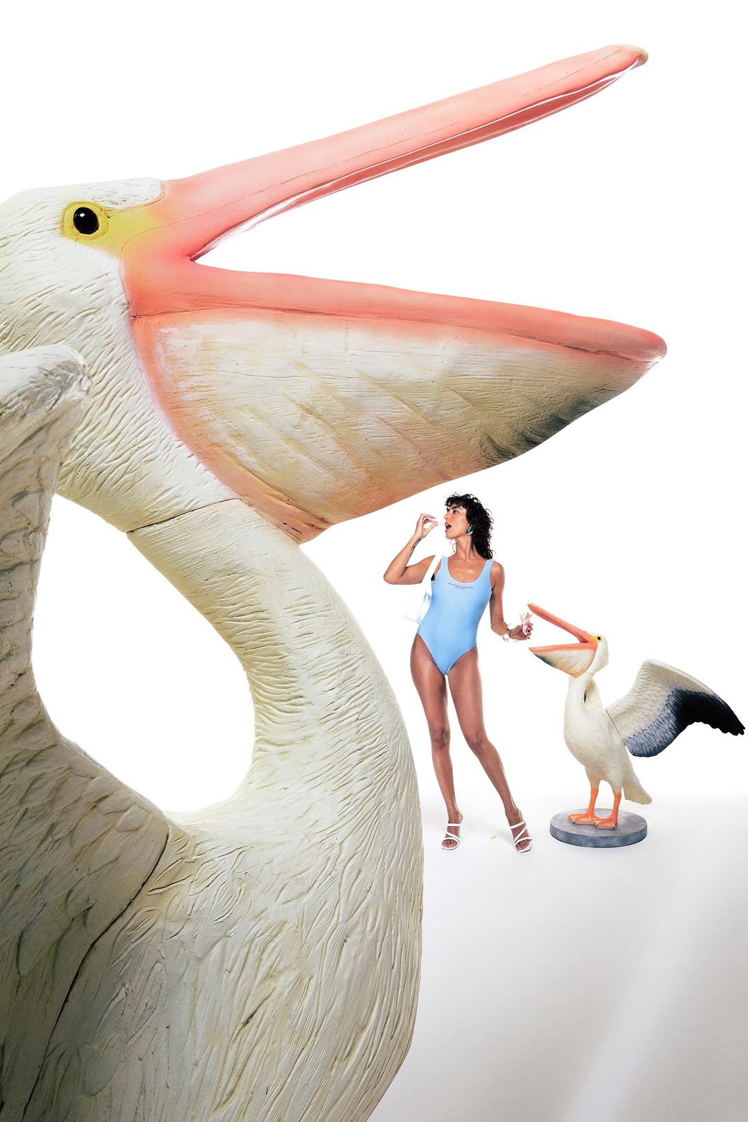 Woman in blue swimsuit stands near oversized pelican statue and another smaller pelican statue while holding a pill in her fingers.