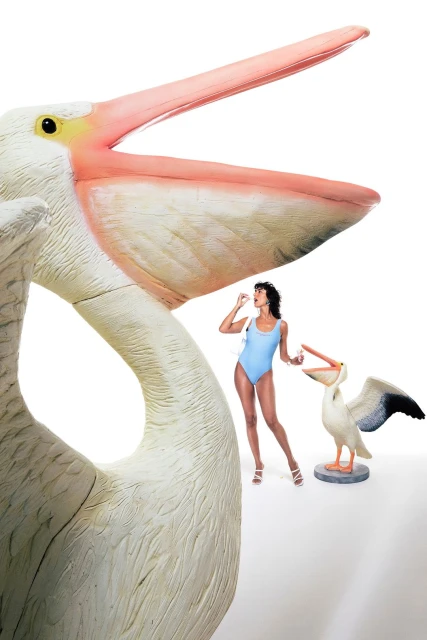 Woman in blue swimsuit stands near oversized pelican statue and another smaller pelican statue while holding a pill in her fingers.