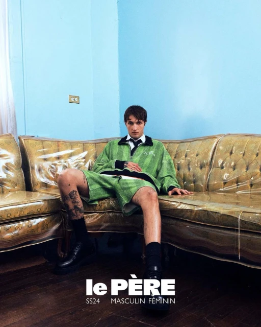 A stylish young adult male poses on a gold couch wearing a green outfit, black socks and shoes inside a room with blue walls.