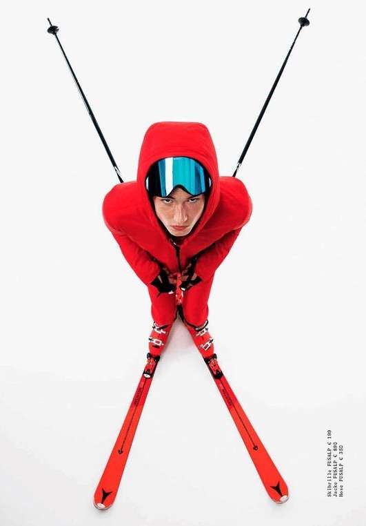 A person in a red ski suit and blue goggles stands on red skis, holding ski poles, viewed from above against a bright white background.