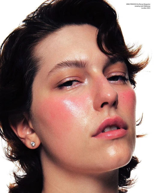 Close-up studio portrait of a young adult woman with short dark hair, fair skin, and pink blush wearing a small stud earring.