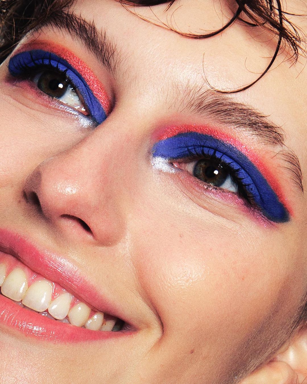 Close-up of a smiling woman with striking blue and coral makeup on her eyelids, enhancing her eyes, set against a soft skin tone.