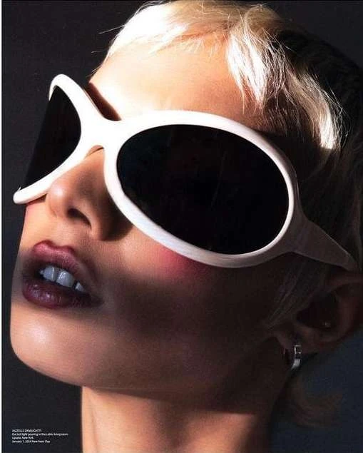 Close-up of a woman with short blonde hair wearing oversized white sunglasses in a studio with dark gray backdrop.
