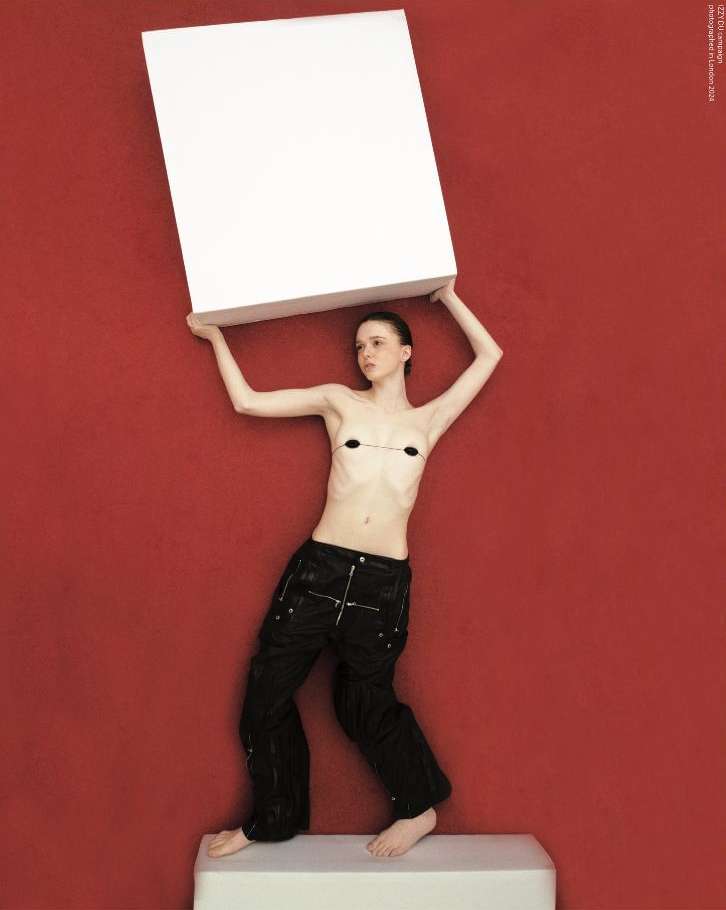 A slender woman stands indoors holding a large white box, clad in dark trousers against a solid red backdrop, balanced on a white cube.