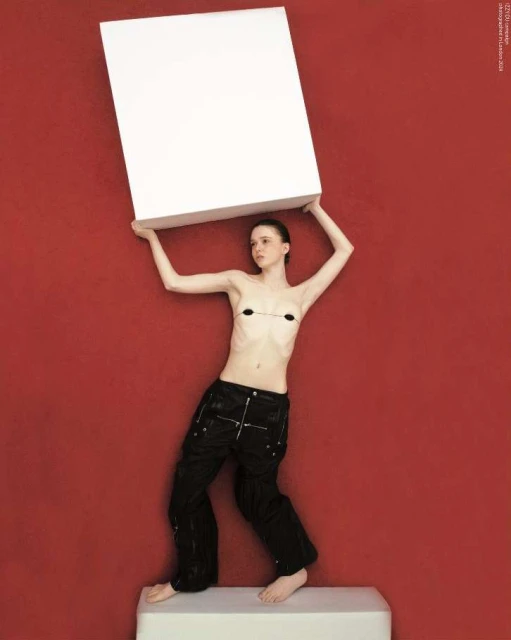 A slender woman stands indoors holding a large white box, clad in dark trousers against a solid red backdrop, balanced on a white cube.
