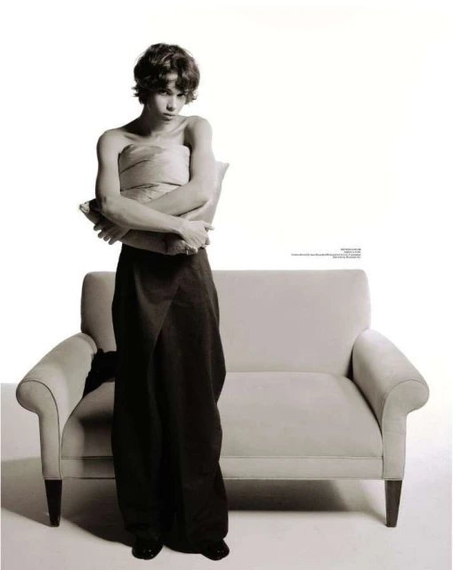 Portrait of a young adult wearing a brown wrap around the waist and holding a pillow in a photo studio setting with a couch.