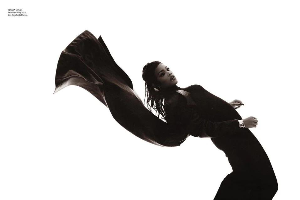 Teyana Taylor poses in a black outfit with a cape-like fabric against a white background in a fashion editorial shot.