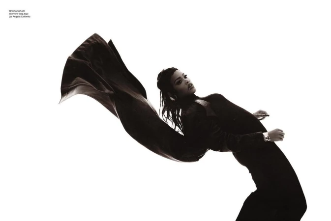 Teyana Taylor poses in a black outfit with a cape-like fabric against a white background in a fashion editorial shot.