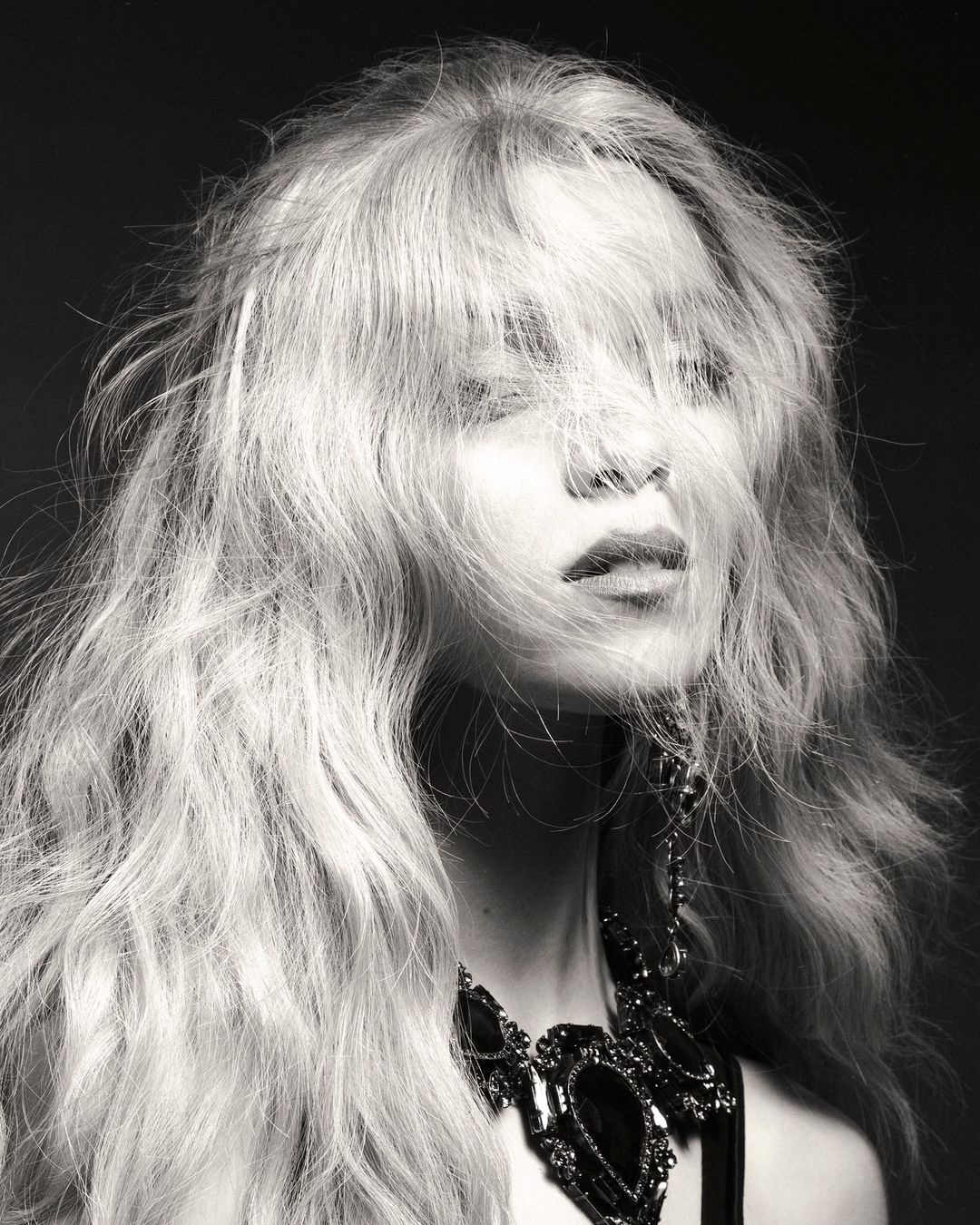 Monochrome portrait of a woman with long tousled hair falling over her eyes wearing a dark ornate necklace.