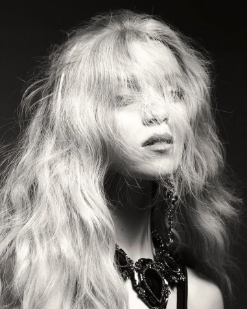 Monochrome portrait of a woman with long tousled hair falling over her eyes wearing a dark ornate necklace.
