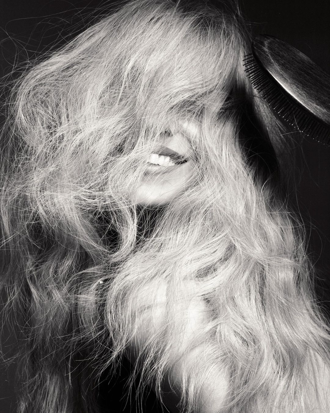 Black and white portrait of a smiling adult female with long, wavy hair partially covering her face, and a hairbrush above.