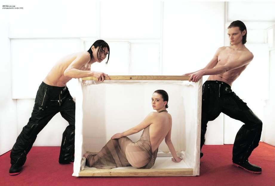 Fashion editorial featuring a woman in a sheer dress inside a box held by two shirtless men against a white backdrop with a red floor.