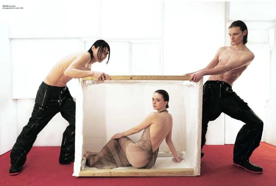 Fashion editorial featuring a woman in a sheer dress inside a box held by two shirtless men against a white backdrop with a red floor.