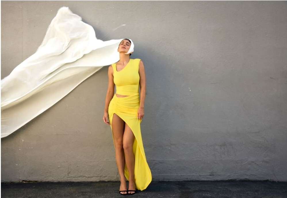 A woman models a yellow dress with a large white veil blowing in the wind against a grey wall in an outdoor fashion shot.