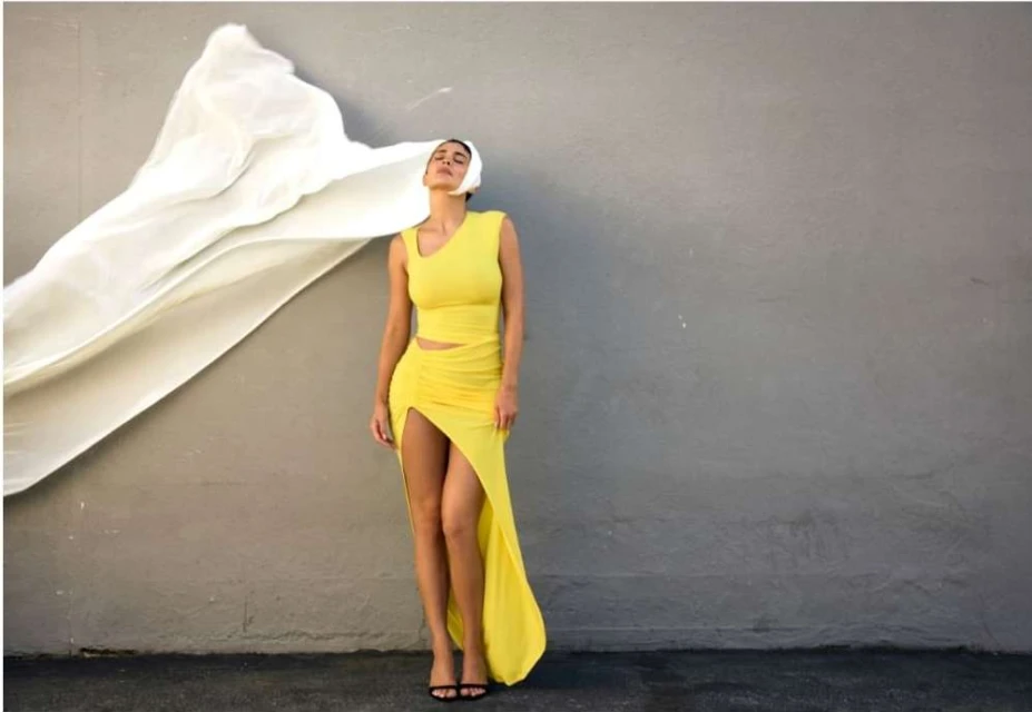 A woman models a yellow dress with a large white veil blowing in the wind against a grey wall in an outdoor fashion shot.