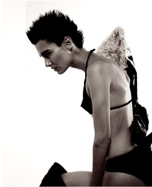 Monochrome fashion shot of a young adult woman with spiky dark hair kneeling with homemade angel wings and bra top against a white backdrop.