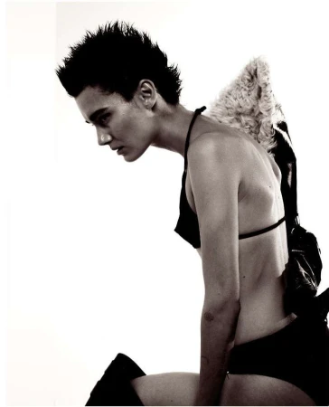 Monochrome fashion shot of a young adult woman with spiky dark hair kneeling with homemade angel wings and bra top against a white backdrop.