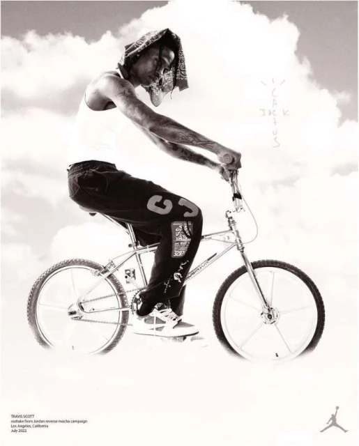 Black and white shot of Travis Scott riding a bike in front of a cloud background, wearing dark pants, white tank top, bandana on head.