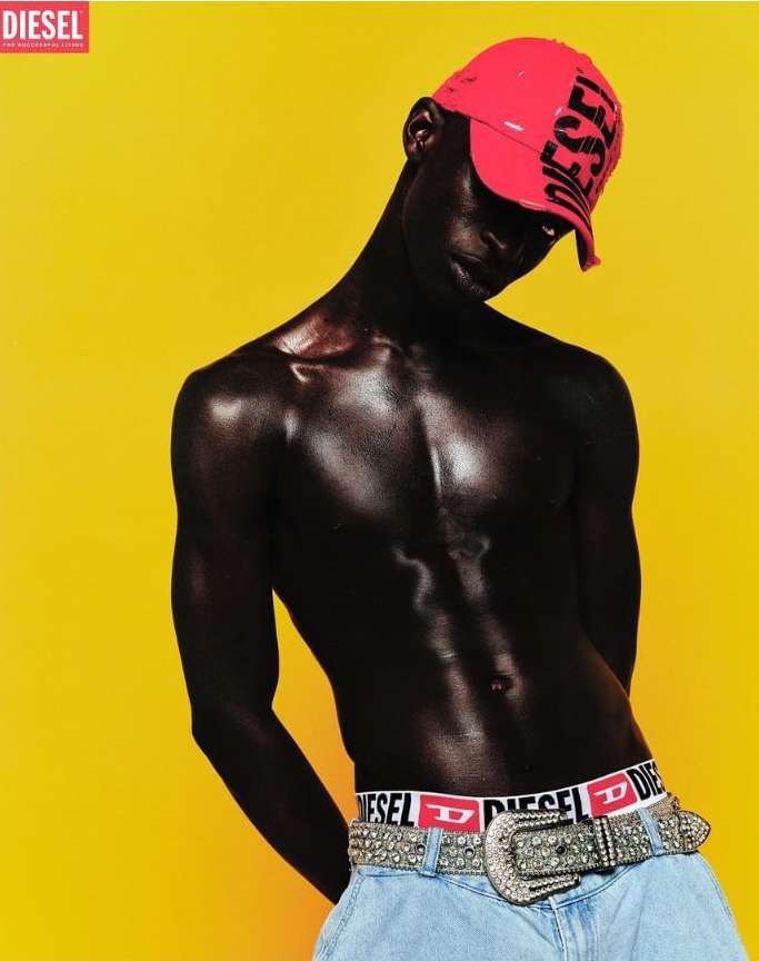 Fashion shot of a dark-skinned man in a red cap and light jeans with a Diesel belt against a bright yellow background in a studio setting.