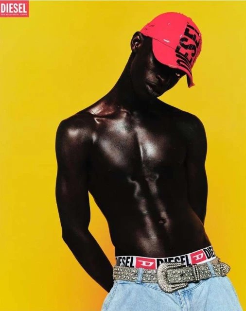 Fashion shot of a dark-skinned man in a red cap and light jeans with a Diesel belt against a bright yellow background in a studio setting.