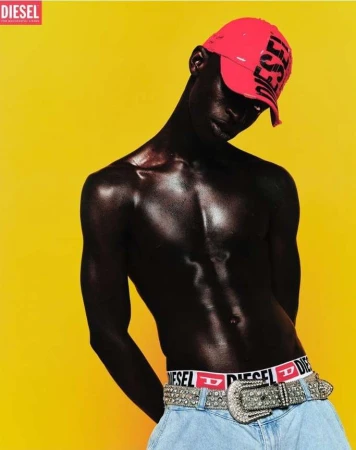 Fashion shot of a dark-skinned man in a red cap and light jeans with a Diesel belt against a bright yellow background in a studio setting.