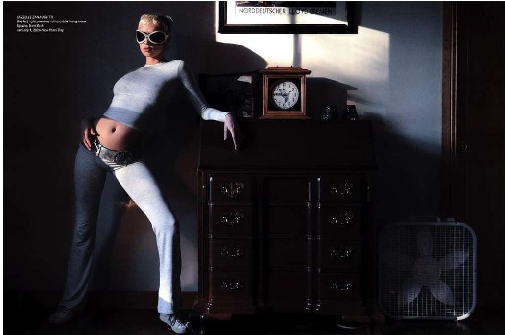 Woman in grey loungewear and sunglasses poses leaning against a wood dresser next to an electric fan in room with sunlight.