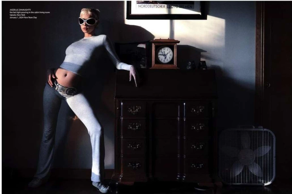 Woman in grey loungewear and sunglasses poses leaning against a wood dresser next to an electric fan in room with sunlight.