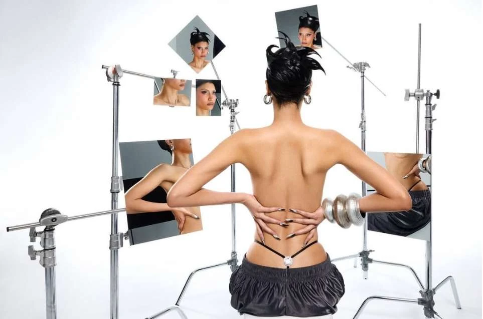 Back view of a slender woman with a sleek updo, wearing black shorts and bracelets, reflected in multiple mirrors against a white backdrop.