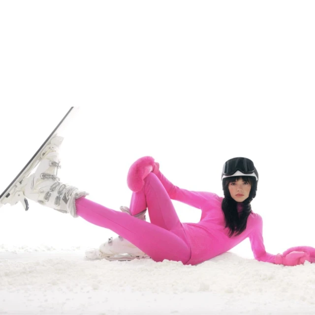 Woman in pink ski suit poses on snow with ski boots and snowboard. Goggles on head. White background. Studio shot. Fashion and sport.