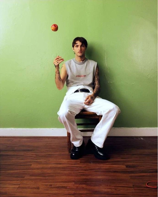 Young adult male with tattoos poses while sitting on a wooden stool indoors, a peach floats above his hand in front of a green wall.