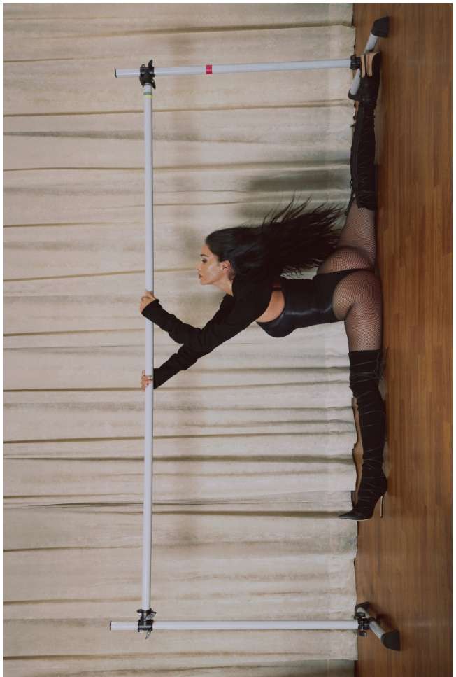 A woman in a black bodysuit and heeled boots performs a split between two vertical metal poles against a background of textured curtains.
