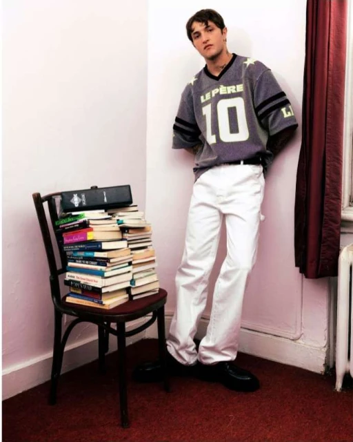 A young man in a jersey and white pants leans against a wall next to a chair piled with books in a room with red carpet.