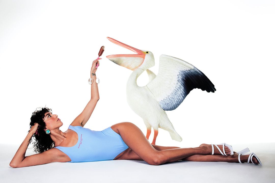 Woman in swimsuit lying next to a decorative pelican, holding an ice cream bar, with a white seamless background in a studio.