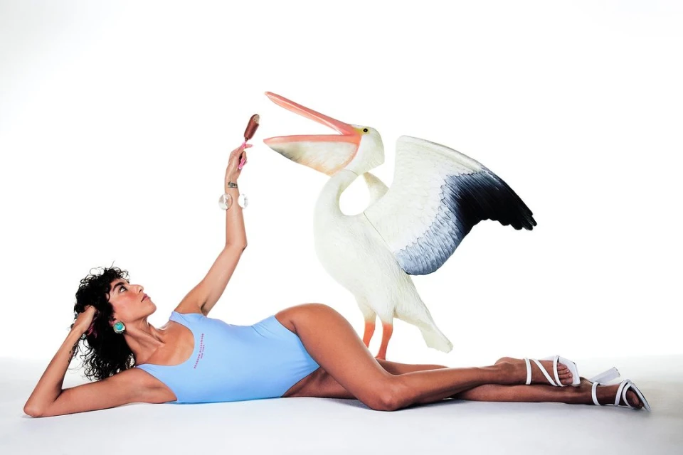 Woman in swimsuit lying next to a decorative pelican, holding an ice cream bar, with a white seamless background in a studio.