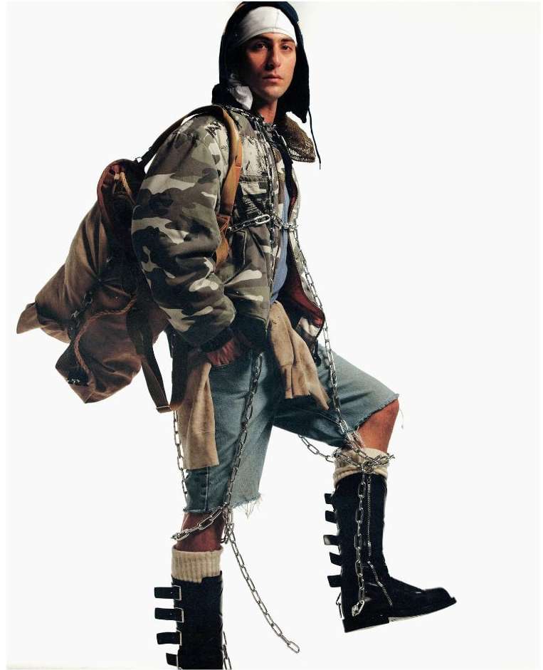 Man in camo jacket, ripped shorts, and black boots with chains, posing in a studio with a backpack, creating a unique and edgy fashion statement.