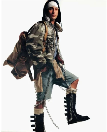 Man in camo jacket, ripped shorts, and black boots with chains, posing in a studio with a backpack, creating a unique and edgy fashion statement.