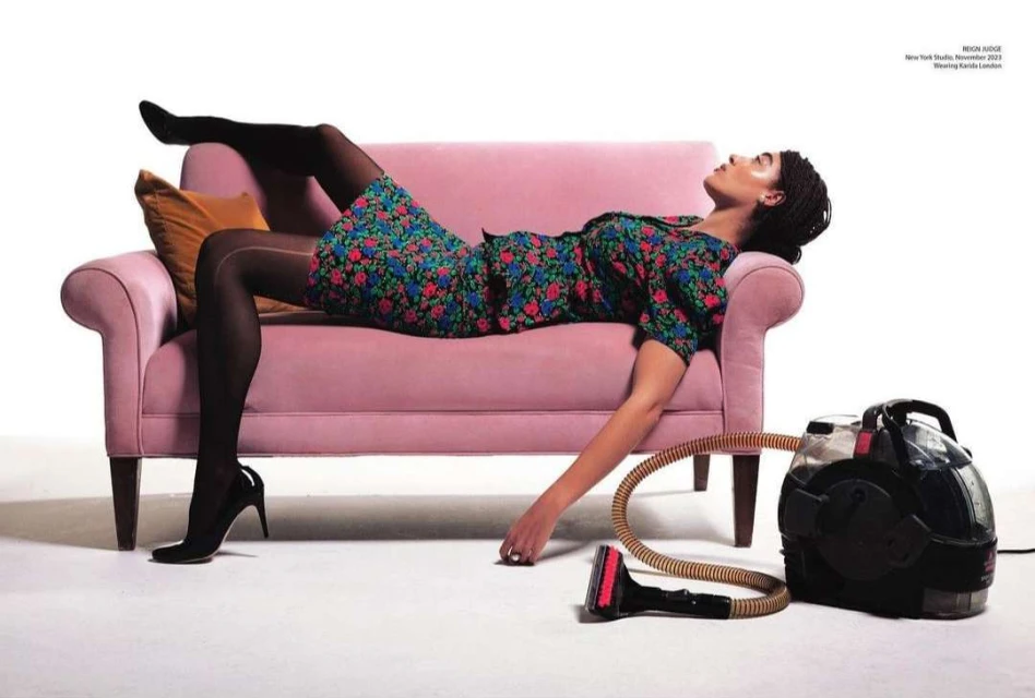 A woman in a floral dress reclines on a pink couch next to a carpet cleaner in a studio setting, showcasing fashion and domesticity.