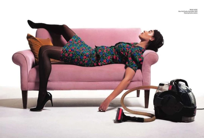 A woman in a floral dress reclines on a pink couch next to a carpet cleaner in a studio setting, showcasing fashion and domesticity.