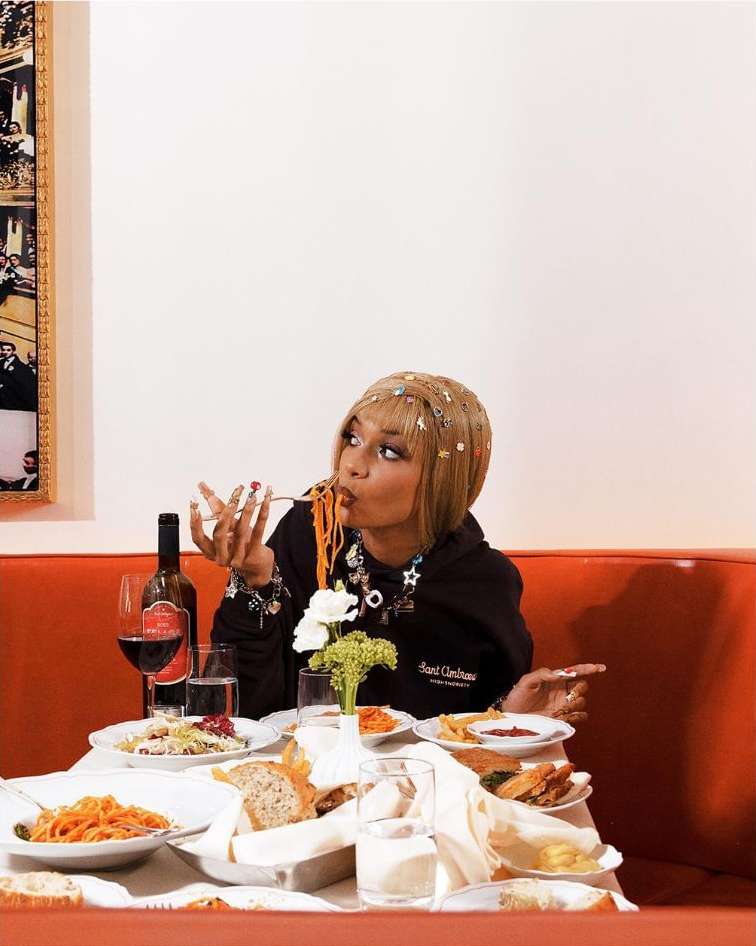 Woman eating spaghetti at a restaurant table with wine, bread, and other food, wearing a black hoodie and accessorized with jewelry.